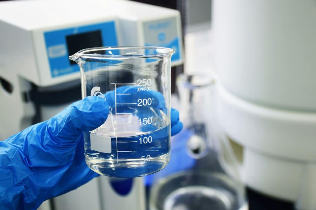 Mão com luva segurando um béquer com amostra de água em laboratório
