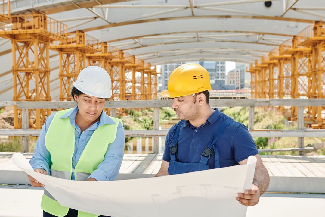 Dois engenheiros com capacete e colete refletivo consultando a planta de uma obra